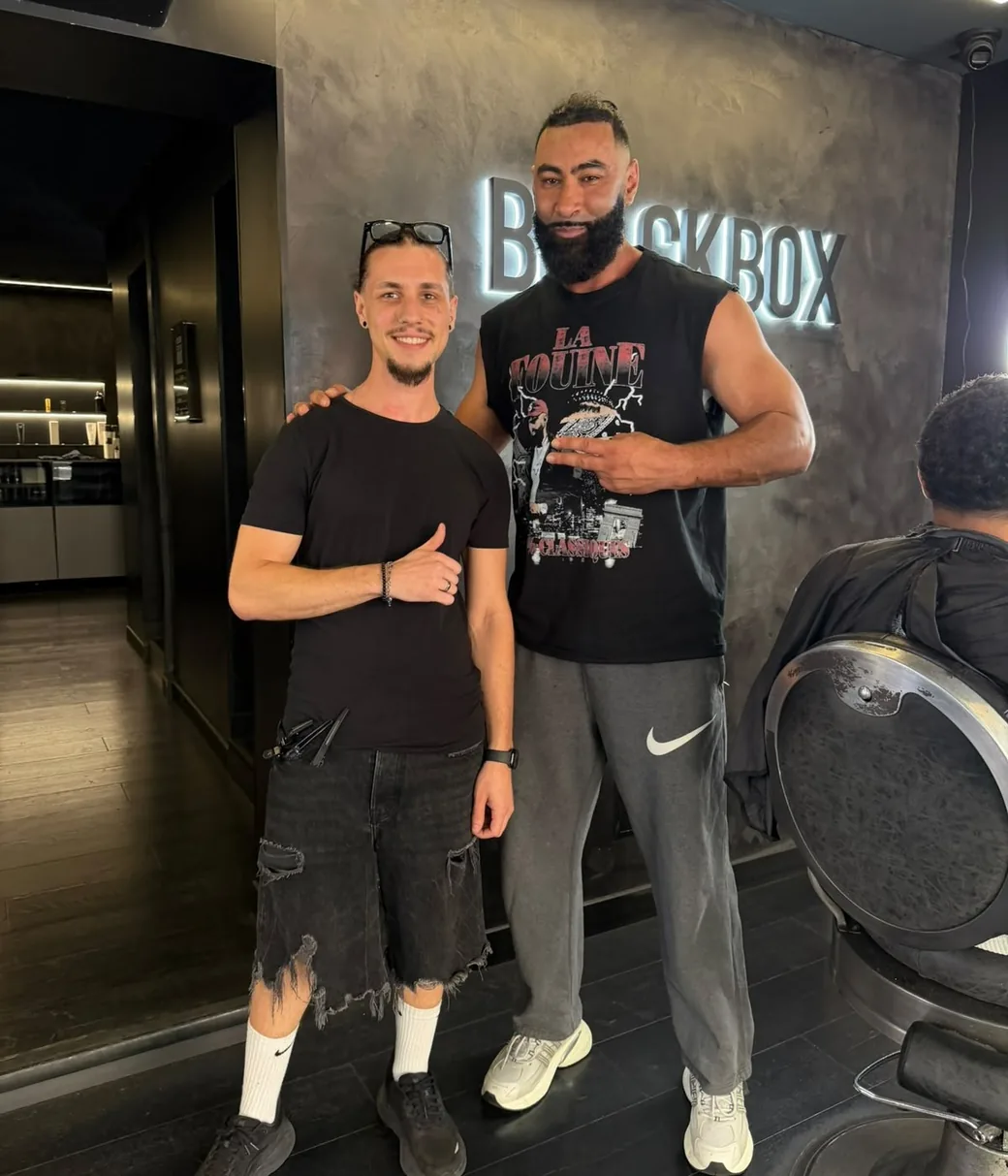 Corentin, ciseaux à la ceinture, aux côtés d’un client de grande taille après sa coupe.