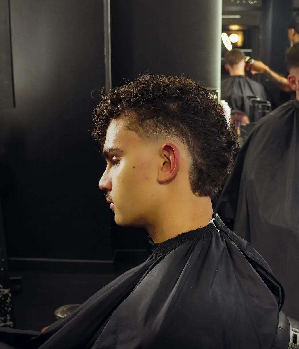 Profil d’un client après la coupe : mulet bouclé, dégradé fondu sur les côtés. Dans le miroir, un autre rendez-vous se termine.